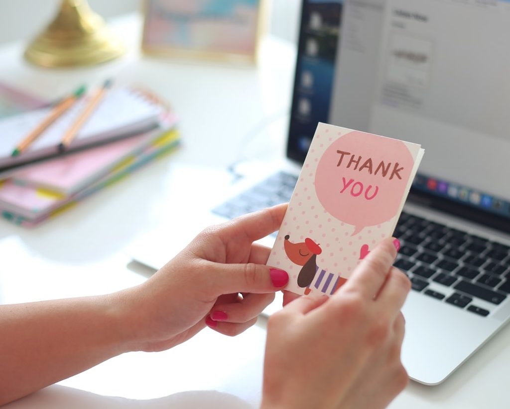 Tangan memegang kartu 'Thank You' dengan ilustrasi lucu, di depan laptop dan buku.