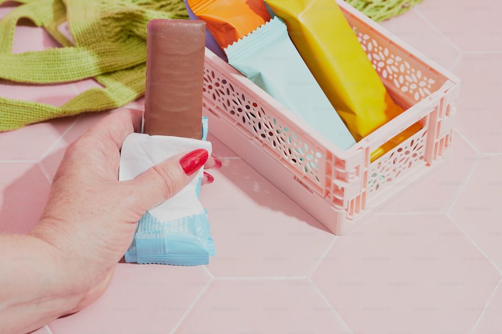 Tangan memegang kemasan makanan di rak plastik dengan warna-warna cerah.