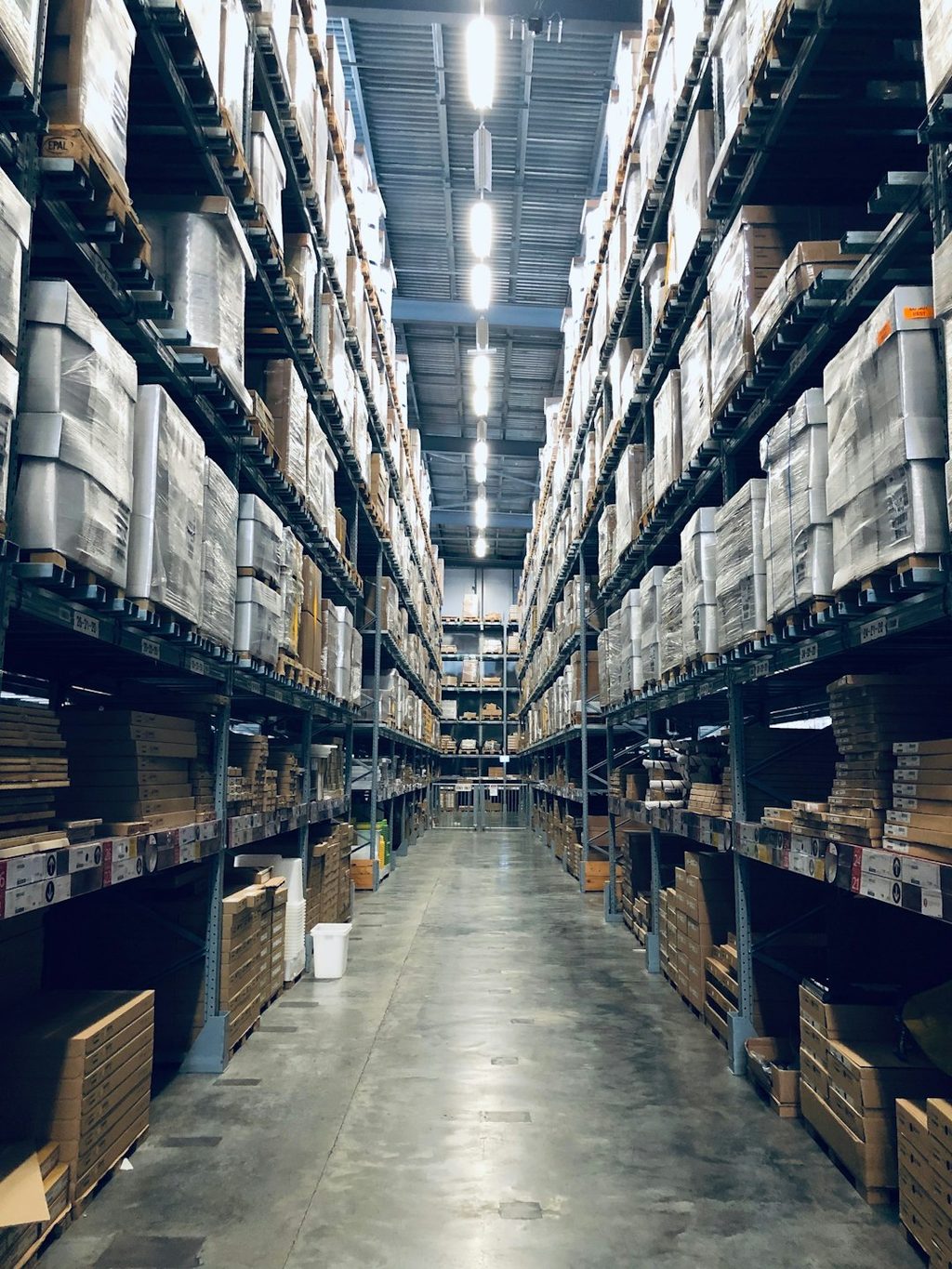 Lorong gudang dengan rak penuh kemasan dan bahan percetakan yang menggambarkan proses kontrol teknis sebelum cetak massal.