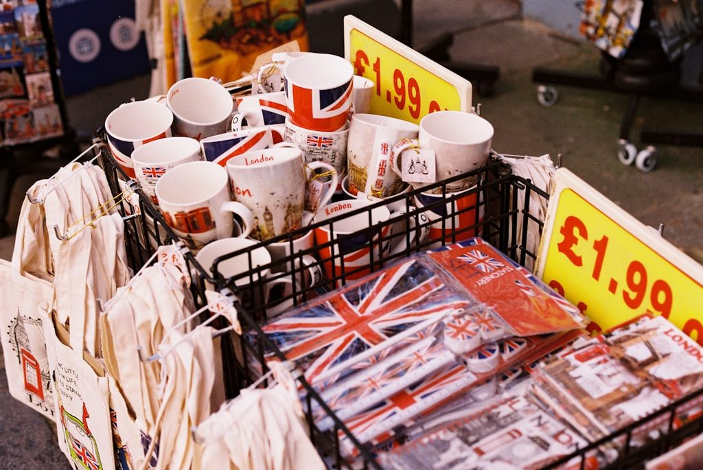 Gambar menampilkan aneka souvenir dan merchandise dengan desain bendera Britania Raya, termasuk mug, kaos, dan aksesori.
