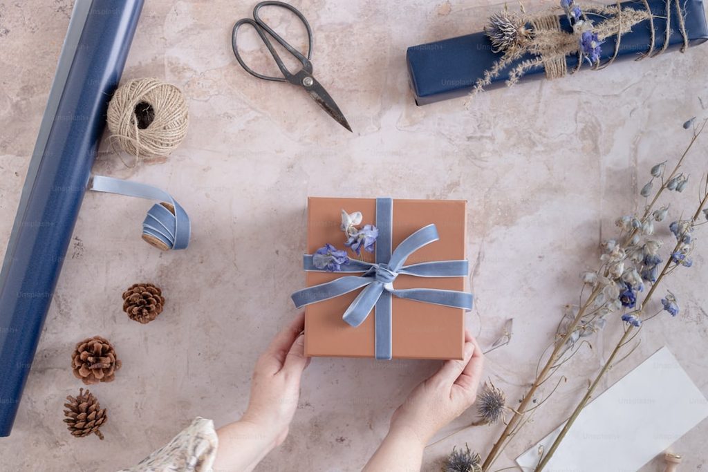 Tangan memegang kotak hadiah kecil dengan tali biru dan bunga dekoratif, di sekelilingnya ada alat pemotong, kertas, dan bunga kering.