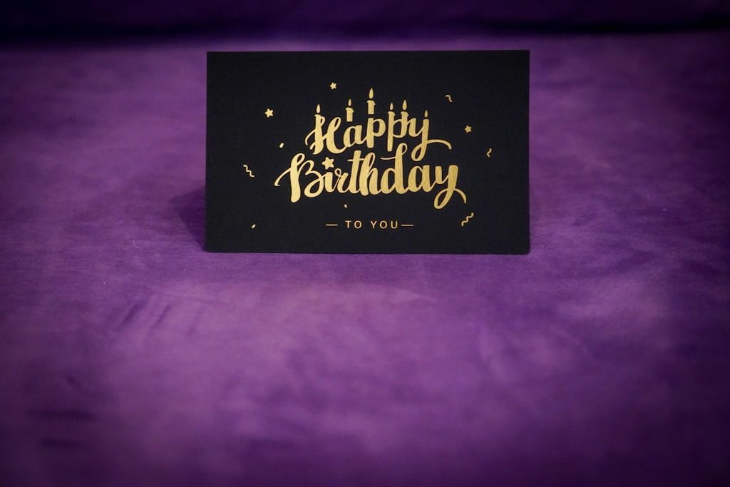 Kartu ucapan Happy Birthday dengan tulisan emas dan hiasan di atasnya, diletakkan di permukaan ungu.