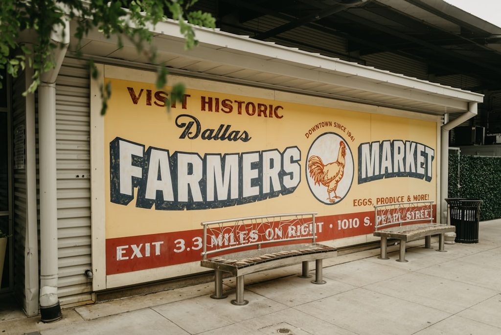 Banner bertuliskan 'Visit Historic Dallas Farmers Market' di depan toko percetakan.