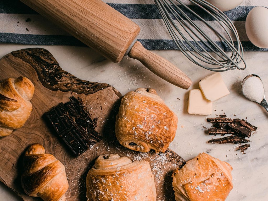 Gambar menunjukkan berbagai jenis roti dan bahan-bahan di atas meja, termasuk telur, gula, dan cokelat.