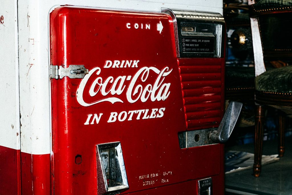 Gambar mesin penjualan koin Coca-Cola merah tua dengan logo dan tagline 'Drink Coca-Cola in Bottles'.