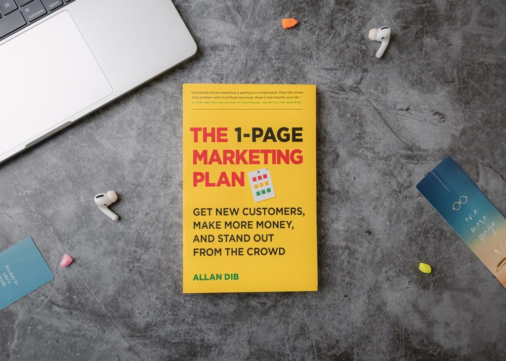Brosur The 1-Page Marketing Plan oleh Allan Dib di meja kerja.