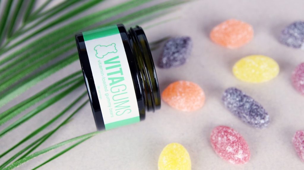 Kemasan botol hijau dengan label 'VITAGUMS' dan permen jelly warna-warni di sekitarnya.