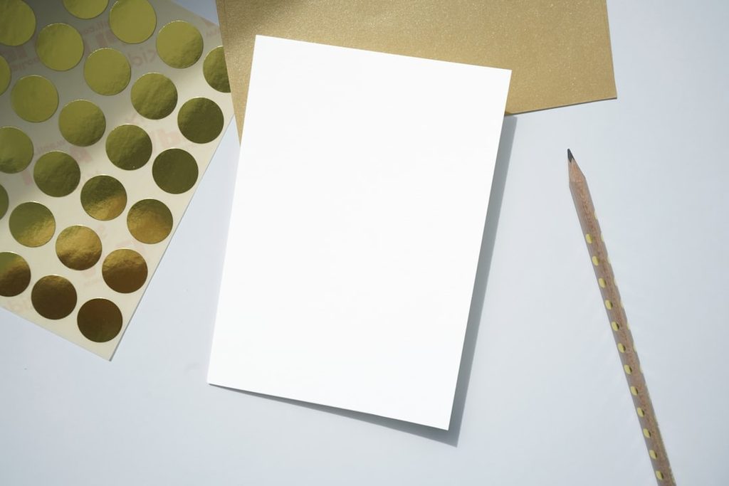 Kertas putih bersih di samping lembar kertas kuning dan pensil kayu untuk ilustrasi persiapan file booklet sebelum cetak.