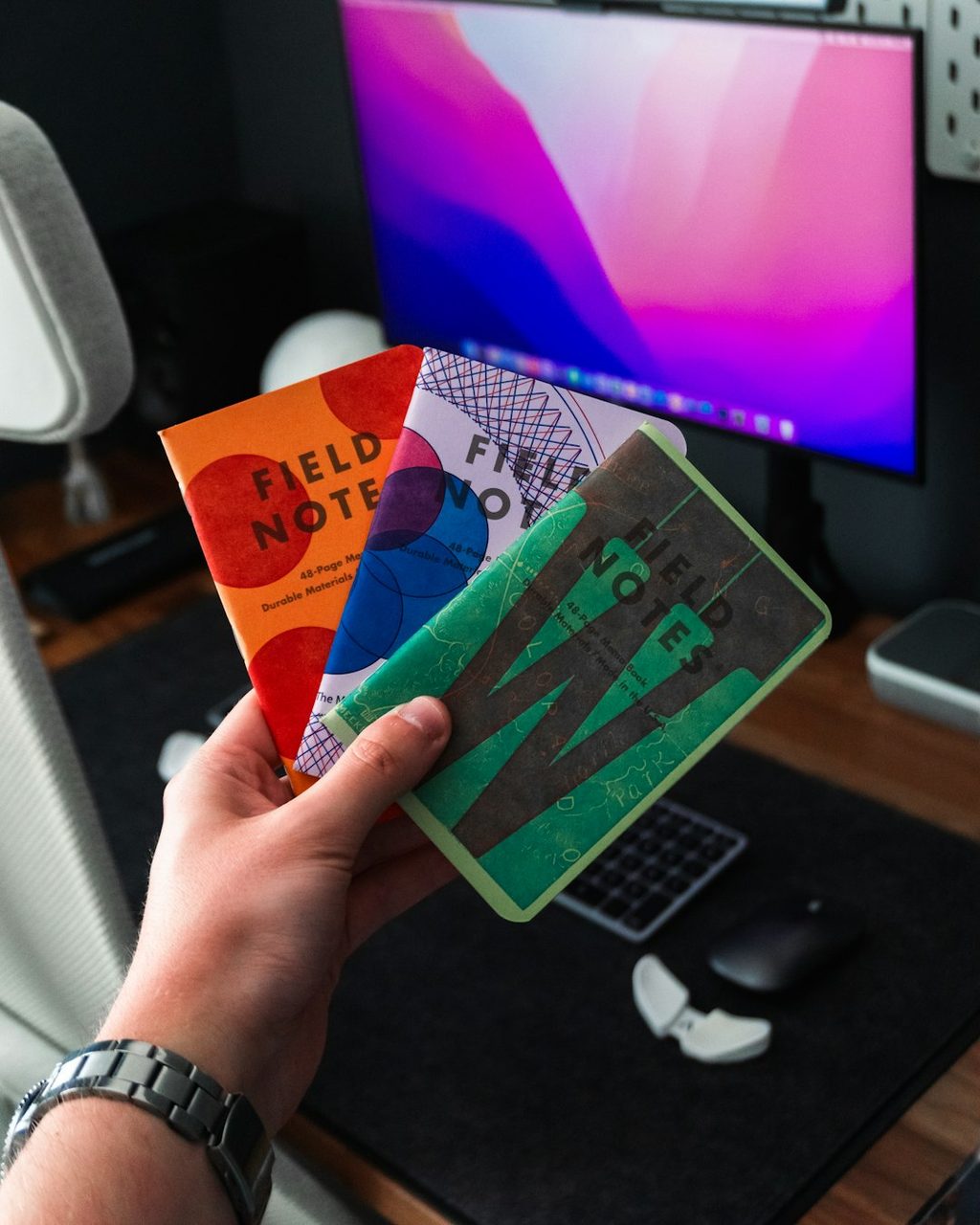 Tangan memegang dua buku Field Notes dengan desain unik di depan monitor komputer.