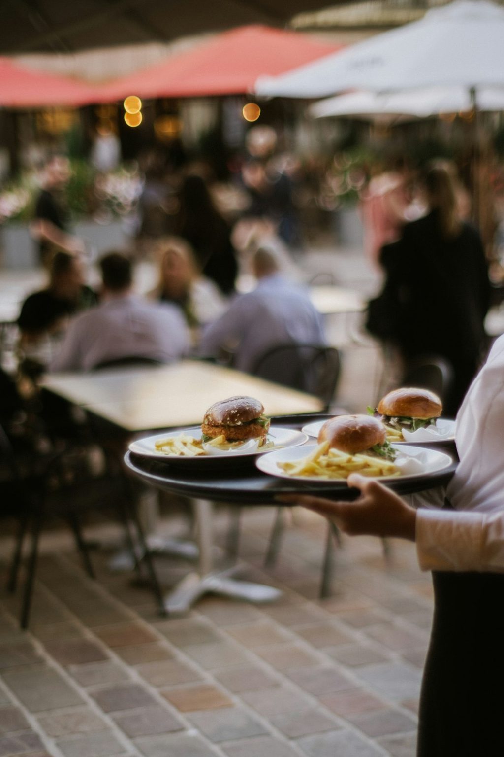 Waiter carrying a tray with burgers and fries in an outdoor cafe setting.