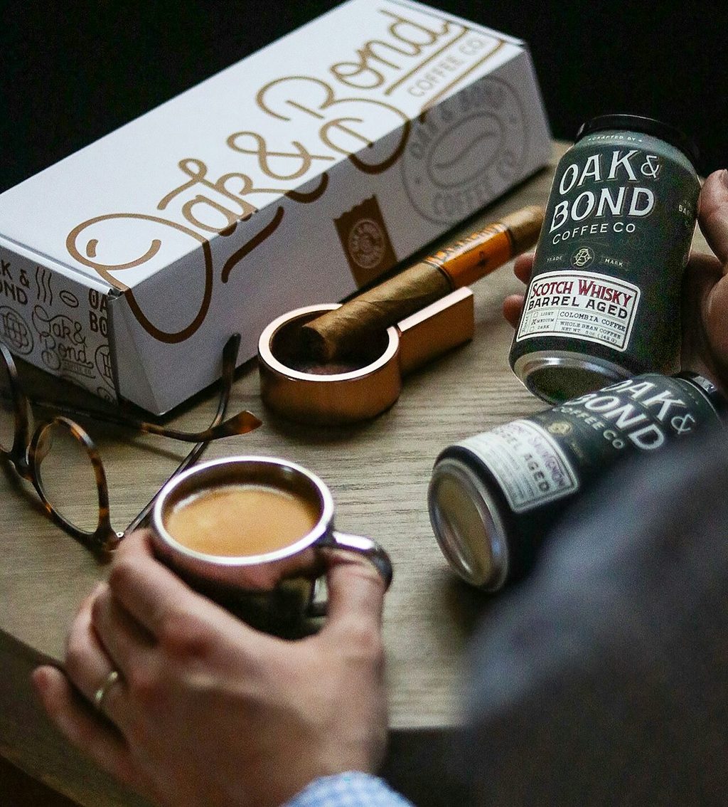 Tangan memegang kopi dan botol minuman Scotch Whisky Barrel Aged dari Oak & Bond Coffee di depan kotak kemasan mereka.