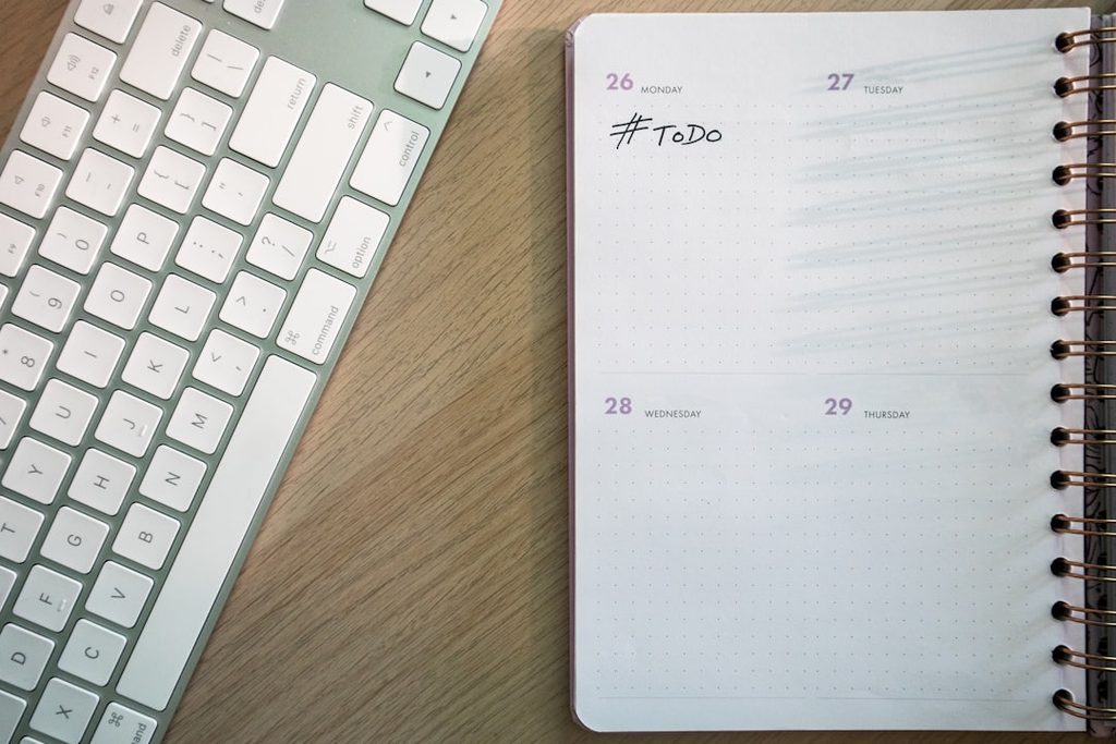Keyboard putih modern dan agenda harian dengan tanda '#ToDo' di meja kayu.