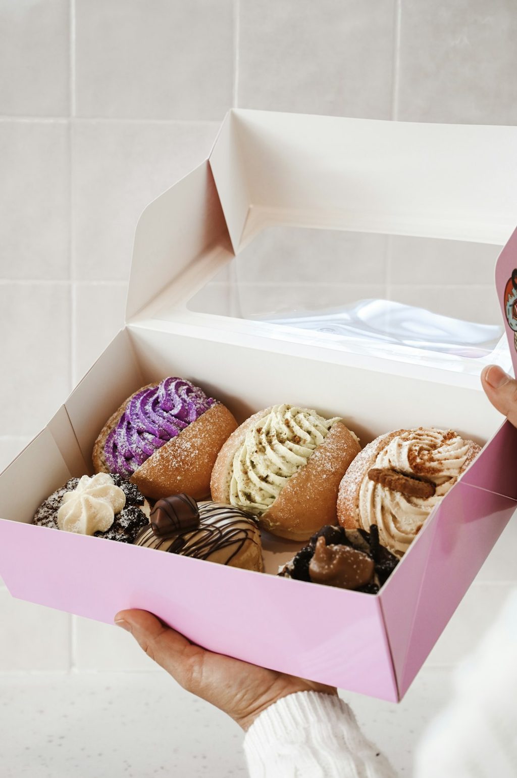 Tangan memegang kotak donat warna pink dengan berbagai jenis donat.