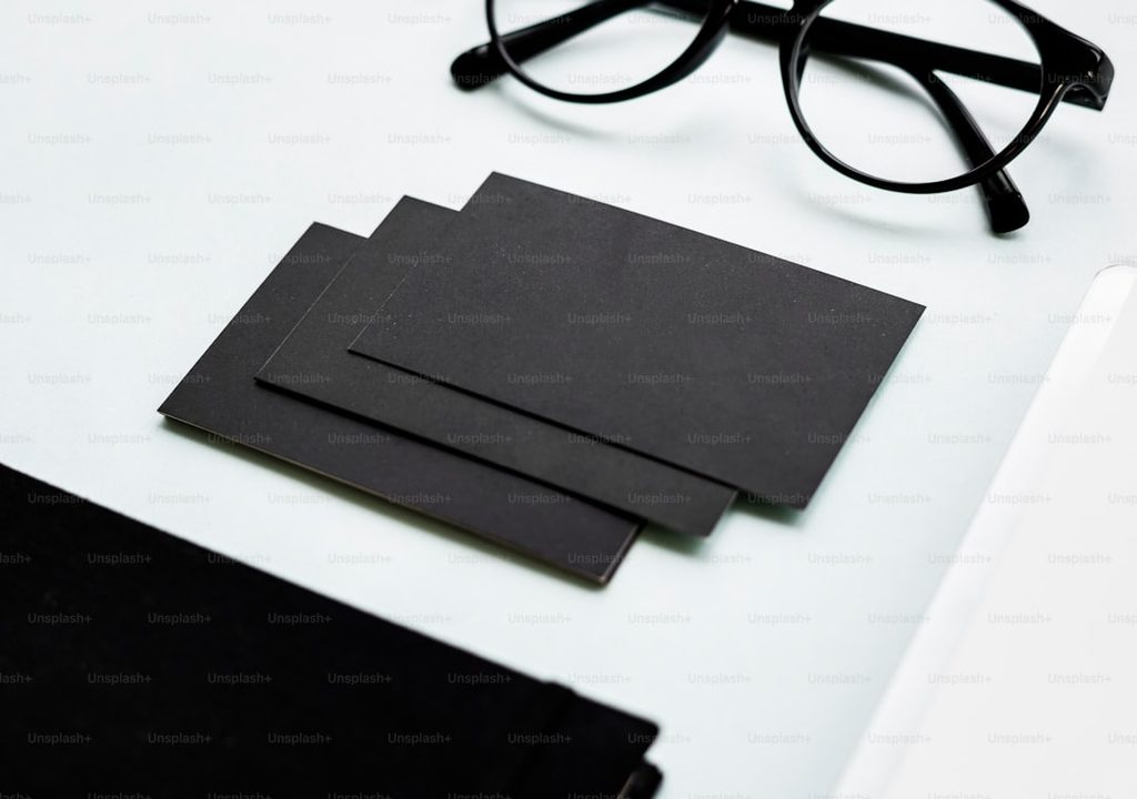 Gambarnya menunjukkan beberapa lembar kartu nama hitam bersih di atas meja putih dengan frame kacamata dan catatan.
