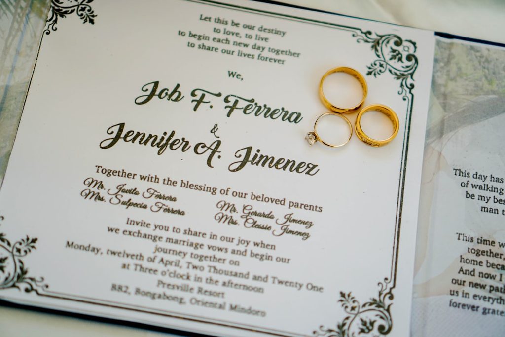 Gambar menunjukkan undangan pernikahan elegan dengan cincin emas dan tanda tangan pengantin.