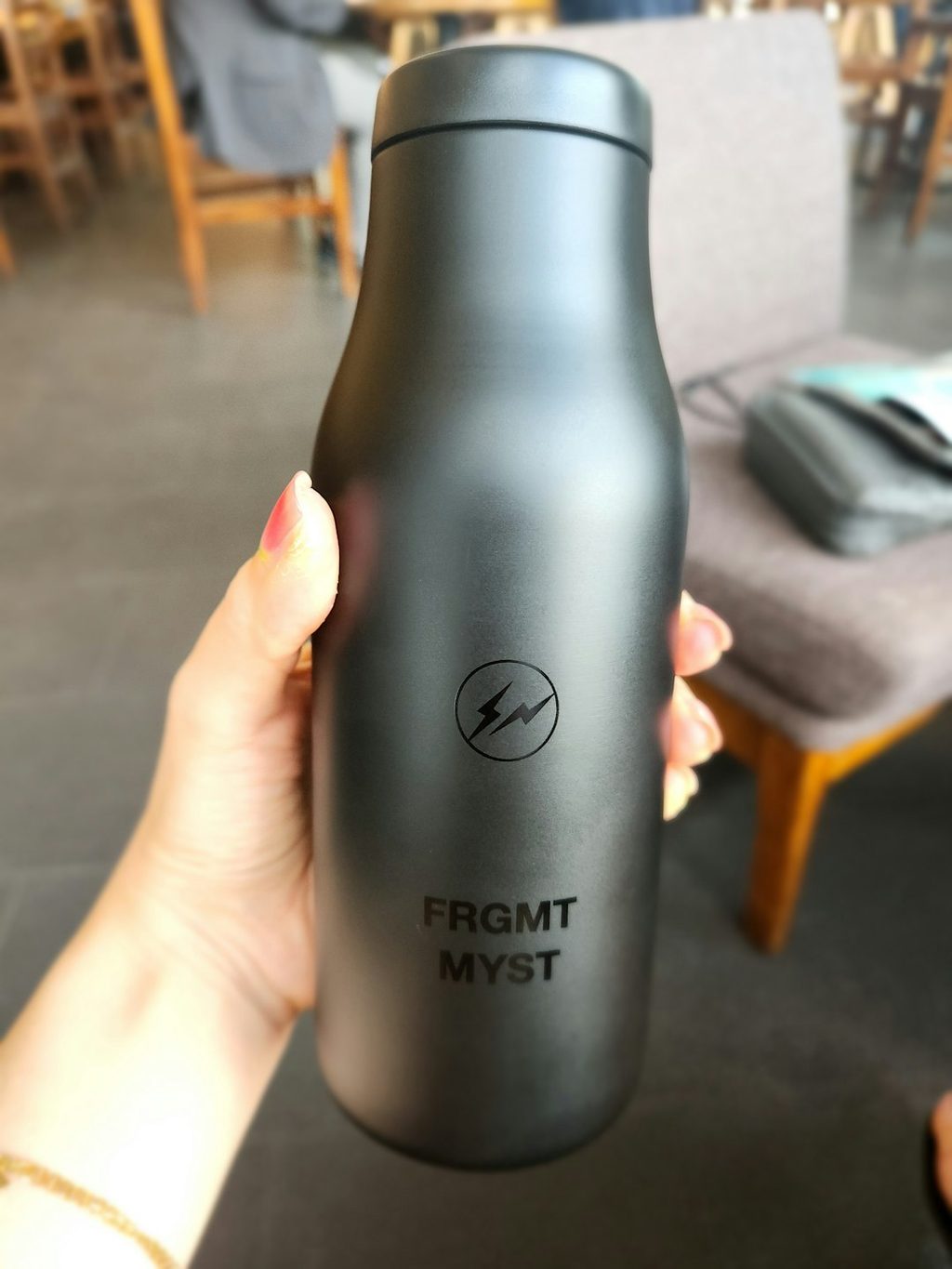 Botol minum hitam dengan logo 'FRGMT MYST' dan simbol listrik, dipegang oleh tangan wanita.