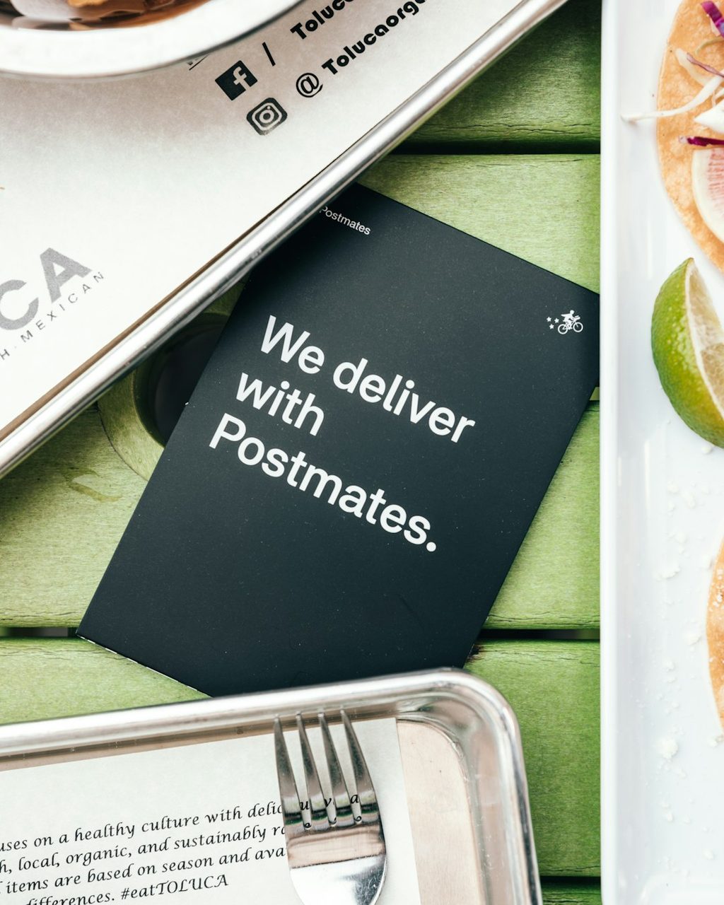 Brosur hitam dengan pesan 'We deliver with Postmates' di atas makanan dan peralatan makan.