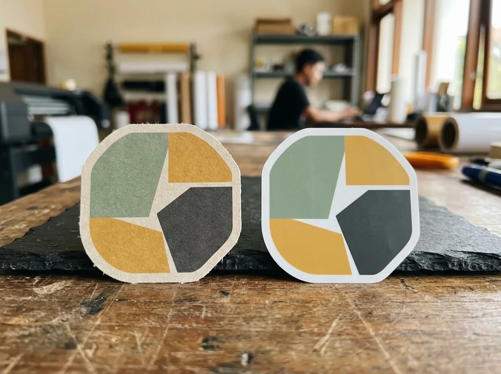 Perbandingan stiker kromo berbahan kertas dan stiker vinyl sintetis tahan air di meja percetakan.