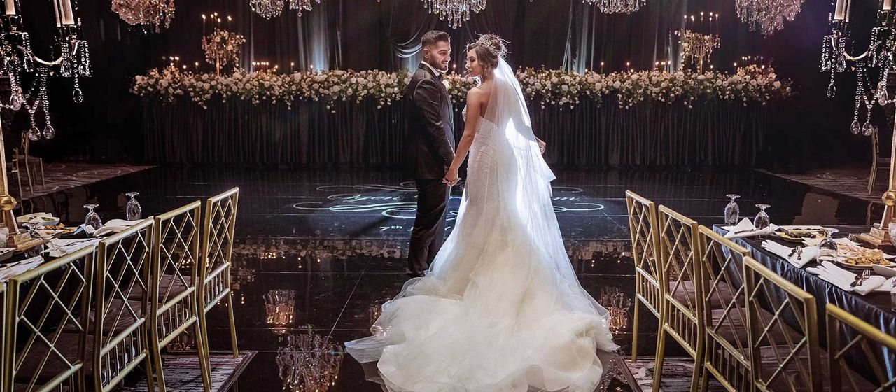 Pasangan pengantin menari di ruang mewah dengan dekorasi bunga dan lampu gantung.