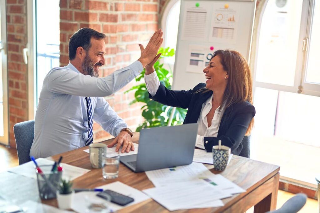 Dua orang sedang memberi high-five di meja kerja, latar ruangan kantor modern.