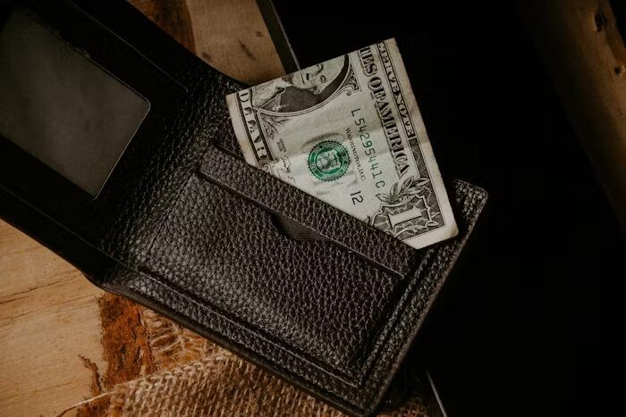 Dompet kulit hitam dengan uang dolar AS di dalamnya, di atas meja kayu.