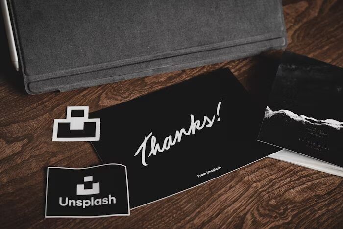 Gambar menunjukkan kartu ucapan 'Thanks!' dari Unsplash dan logo Uprint.id di atas meja kayu.