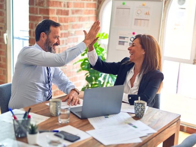 Dua orang sedang memberi high-five di meja kerja, latar ruangan kantor modern.