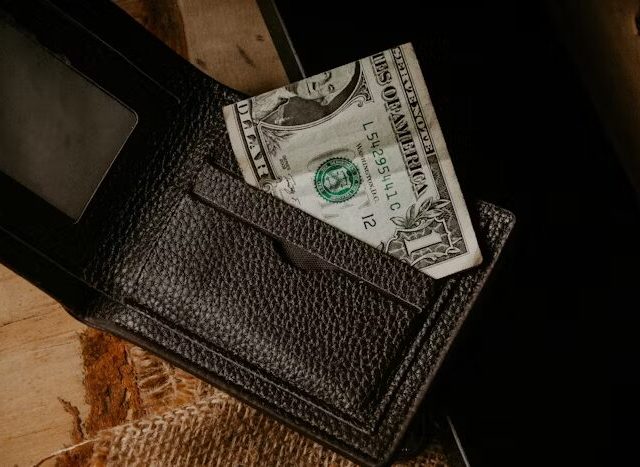 Dompet kulit hitam dengan uang dolar AS di dalamnya, di atas meja kayu.