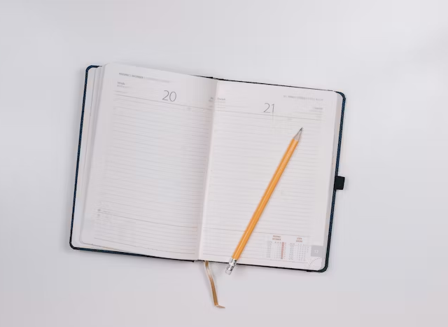 Buku agenda terbuka dengan lembaran kosong dan pensil di sampingnya untuk kebutuhan kerja harian.