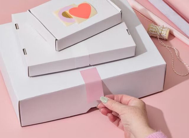 Tumpukan kotak putih dengan stiker hati warna-warni di atasnya, disusun rapi di atas meja berwarna pink.