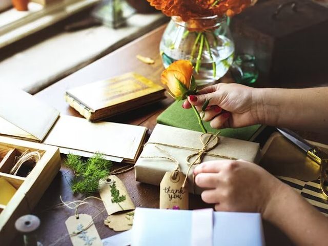 Tangan memegang bunga mawar oranye di atas kotak hadiah yang dikemas dengan kertas kraft dan tali.