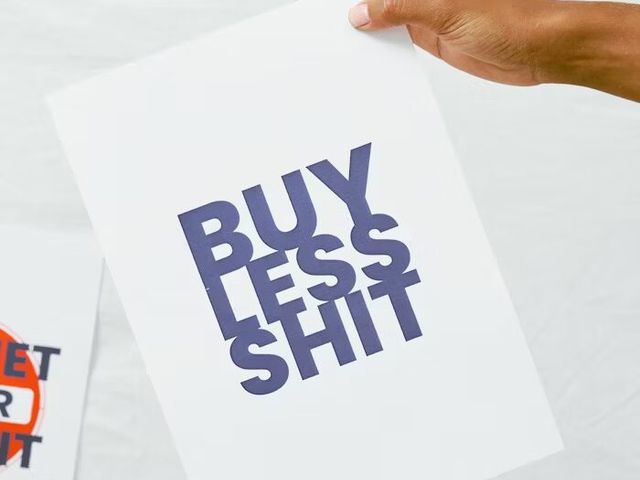 Tangan memegang poster putih dengan pesan 'BUY LESS SHIT' dalam huruf besar.