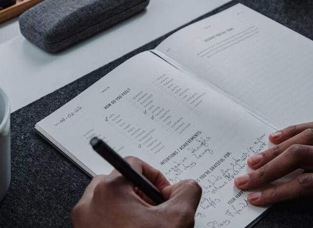 Tangan menulis dalam buku agenda dengan pena di meja kerja.