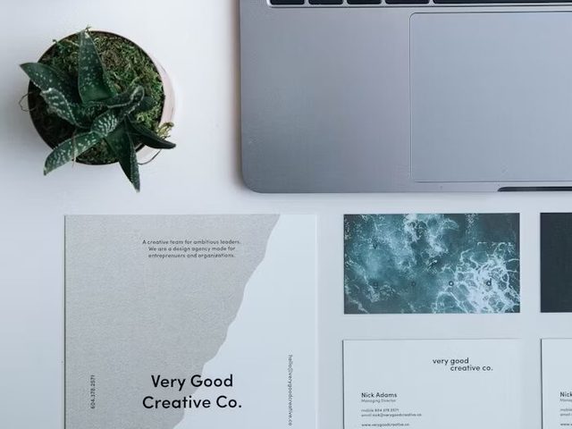 Tampilan kerja studio desain dengan laptop, pot tanaman, dan kartu nama 'Very Good Creative Co.'