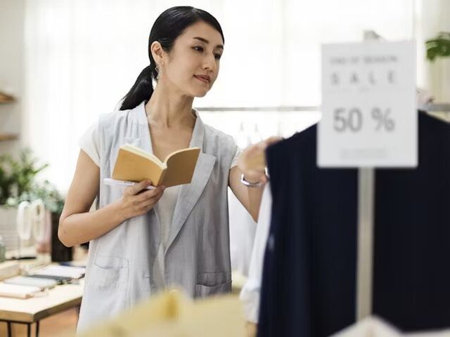 Perempuan membaca buku sambil memilih pakaian di toko dengan penawaran diskon 50%.