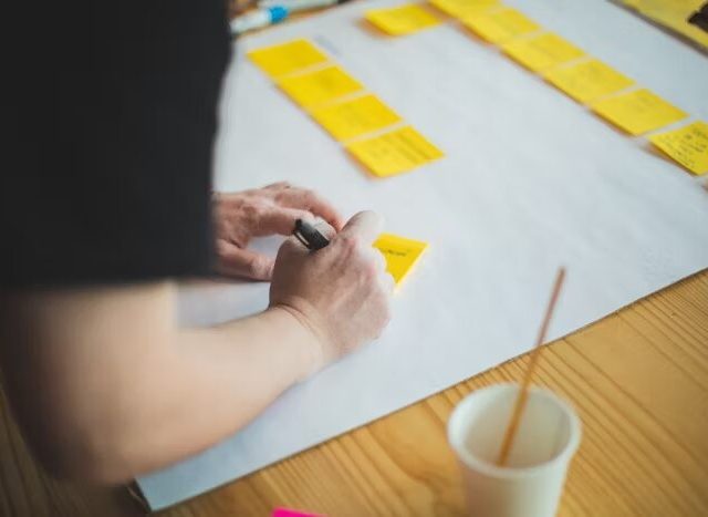 Tangan memegang marker dan menulis di sticky notes, ditempatkan di atas lembaran kertas.