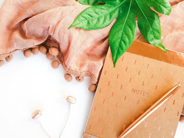 Notebook with rose gold accents and pen on a white background with green leaves and fabric.