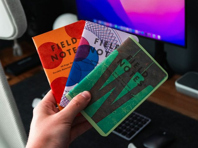 Tangan memegang dua buku Field Notes dengan desain unik di depan monitor komputer.